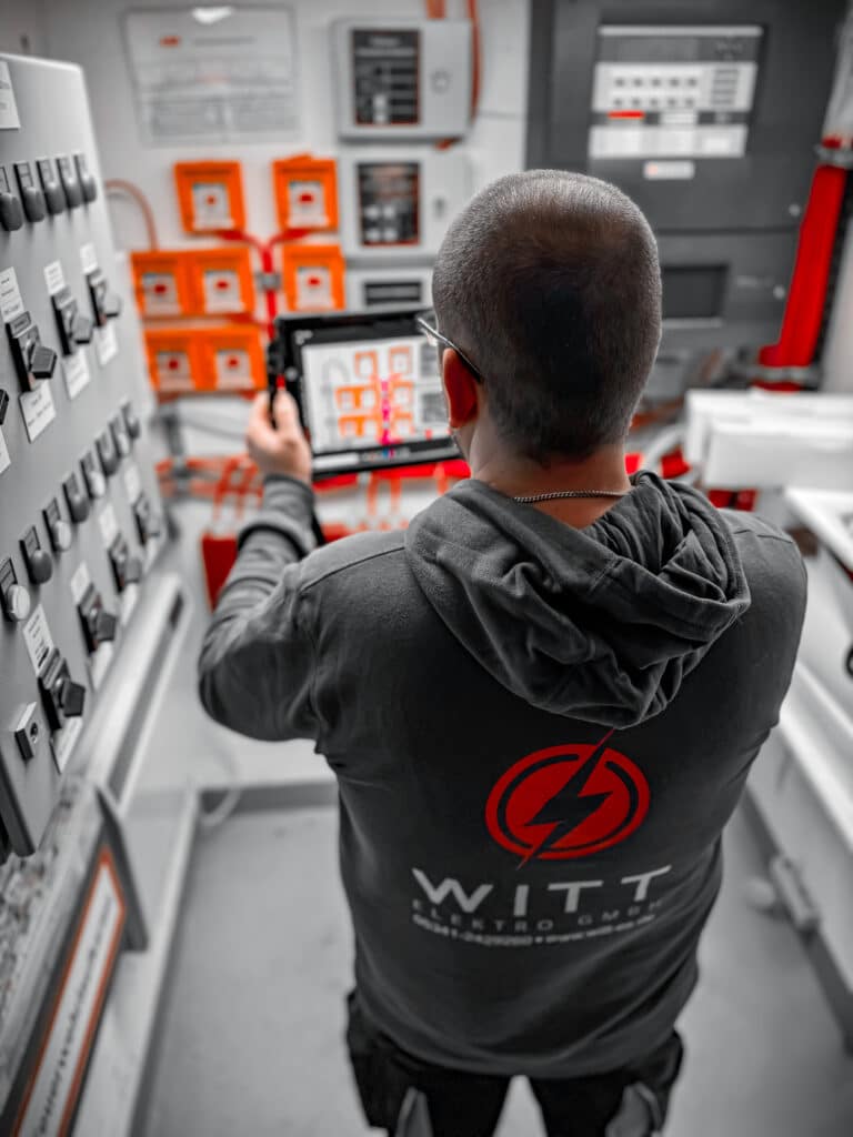 Eine Person mit kurzen Haaren und einem dunklen Kapuzenpullover mit einem roten Logo und Text auf dem Rücken steht in einem Raum voller Schalttafeln und Schalter. Sie hält ein Tablet in der Hand, auf dem ein Aufbau der Brandmeldeanlage Wolfenbüttel angezeigt wird, der dem vor ihnen liegenden ähnelt.