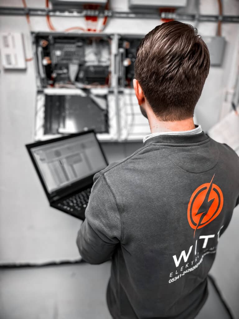 Eine Person mit kurzen Haaren und einem dunkelgrauen Hemd mit einem orangefarbenen Logo steht vor einem offenen Schaltschrank. Sie hält einen Laptop mit sichtbarem Bildschirm in der Hand und arbeitet wahrscheinlich an der Anlage der Brandmeldeanlage Wolfenbüttel oder führt Wartungsarbeiten durch.