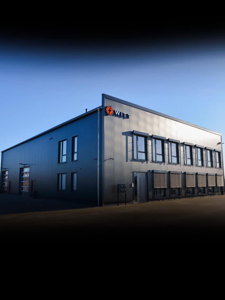 Ein großes, modernes Industriegebäude mit Metallfassade und zwei Fensterebenen beherbergt die Brandmeldeanlage Wolfenbüttel. Die Sonne wirft Schatten auf das Gebäude, während das „WIT“-Logo in der oberen Ecke hervorsticht. Der Himmel bleibt klar und blau.